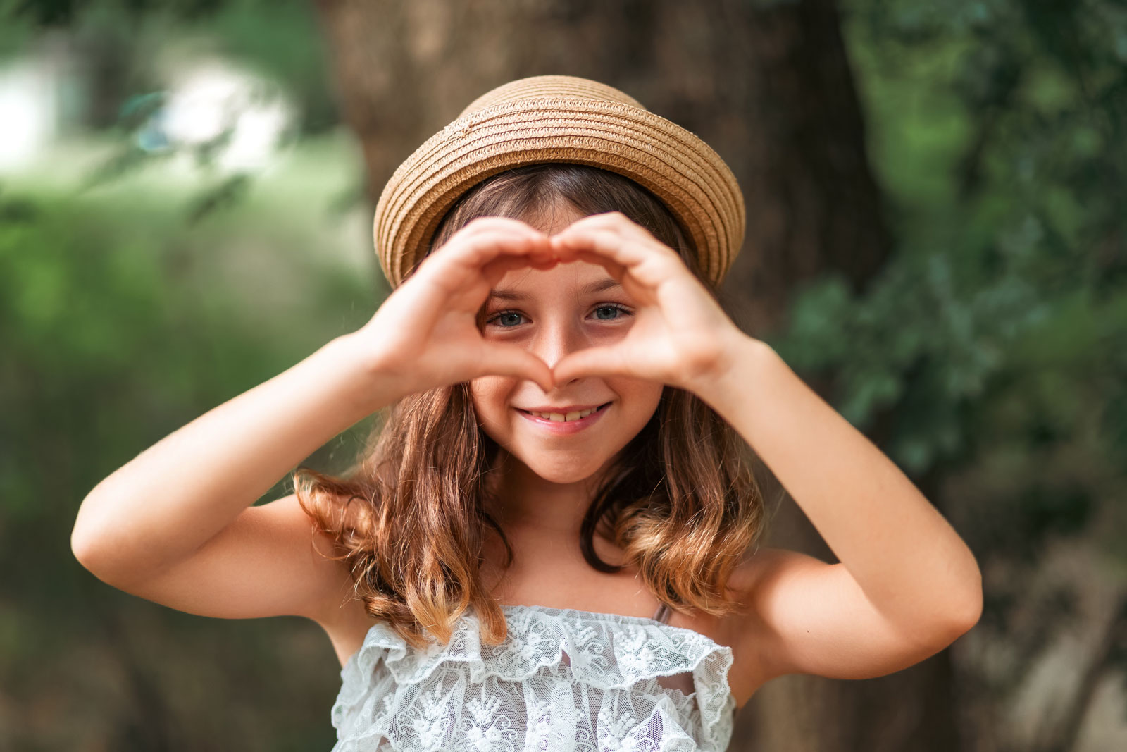 Portrait of a little Caucasian girl in a straw hat showing a heart sign with her fingers. There is a park in the background. Portrait of a little Caucasian girl in a straw hat showing a heart sign with her fingers. There is a park in the background.